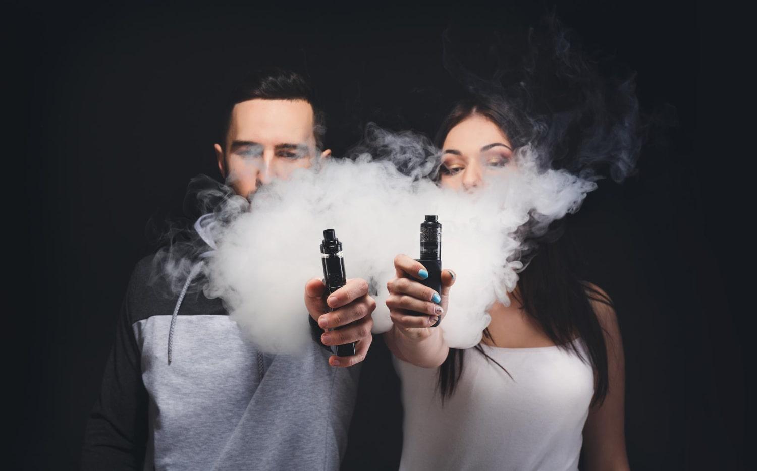 couple vaping unrecognizable young man woman clouds smoke showing their vapes camera black studio background relationship vape addiction concept with copy space min
