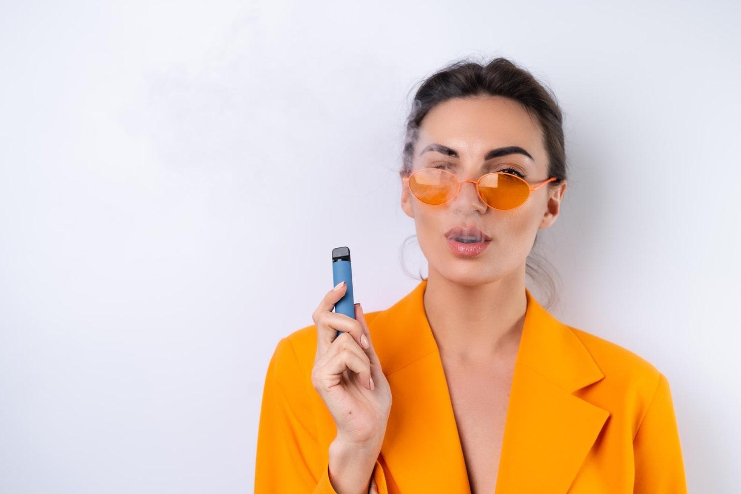 young woman trendy stylish glasses bright orange oversized jacket white background smokes electronic cigarette min